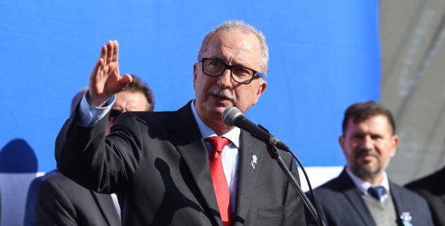 Fotografía del gobernador de Misiones, Hugo Passalacqua, hablando frente a un micrófono durante un acto oficial. Viste traje oscuro y corbata roja, gesticulando con la mano derecha levantada.