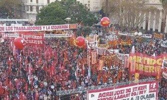 La escena refleja un acto político del Frente de Izquierda Una multitud de personas se encuentra en Plaza de Mayo. Predominan los colores rojo, amarillo y blanco, con numerosas banderas, pancartas y grandes globos rojos flotando sobre el grupo. Se leen letreros con consignas políticas, incluyendo menciones al 'Frente de Izquierda de los Trabajadores', 'Partido Obrero' y 'Izquierda Socialista'.
