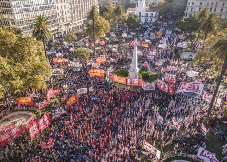 Fotografía aérea de una manifestación masiva en Plaza de Mayo, Buenos Aires. La plaza está colmada de personas con banderas rojas. Se destacan carteles del 'Frente de Izquierda de los Trabajadores' y del 'Partido Obrero'. Hay globos rojos de gran tamaño flotando sobre la multitud y edificios históricos al fondo, como la Catedral Metropolitana.