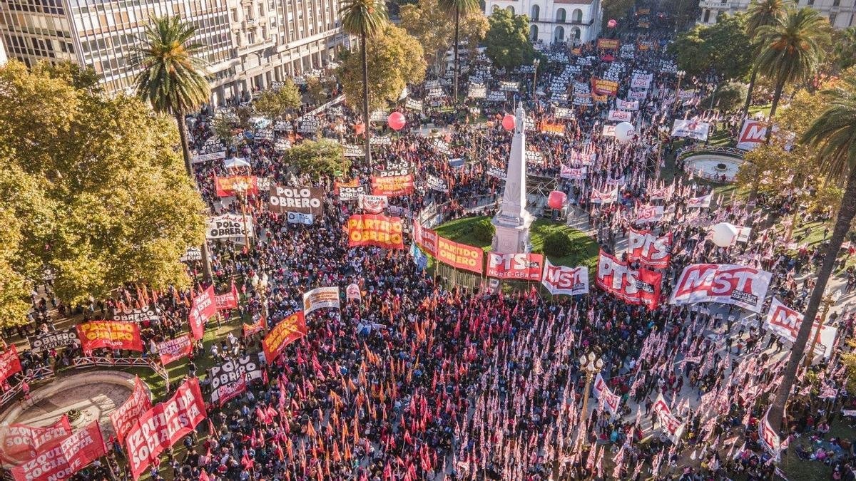 Fotografía aérea de un acto masivo del Frente de Izquierda en Plaza de Mayo, Buenos Aires. La plaza está colmada de personas con banderas rojas. Se destacan carteles del 'Frente de Izquierda de los Trabajadores' y del 'Partido Obrero'. Hay globos rojos de gran tamaño flotando sobre la multitud y edificios históricos al fondo, como la Catedral Metropolitana.
