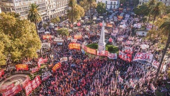 Fotografía aérea de un acto masivo del Frente de Izquierda en Plaza de Mayo, Buenos Aires. La plaza está colmada de personas con banderas rojas. Se destacan carteles del 'Frente de Izquierda de los Trabajadores' y del 'Partido Obrero'. Hay globos rojos de gran tamaño flotando sobre la multitud y edificios históricos al fondo, como la Catedral Metropolitana. Fotografía aérea de un acto masivo del Frente de Izquierda en Plaza de Mayo, Buenos Aires. La plaza está colmada de personas con banderas rojas. Se destacan carteles del 'Frente de Izquierda de los Trabajadores' y del 'Partido Obrero'. Hay globos rojos de gran tamaño flotando sobre la multitud y edificios históricos al fondo, como la Catedral Metropolitana.