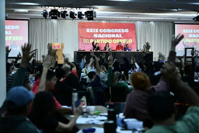 "Vista desde el fondo de un salón lleno de gente donde muchas personas tienen las manos levantadas para votar o participar. Al frente, un panel de cuatro personas está sentado en una mesa larga sobre un escenario. Detrás de ellos hay una gran pancarta roja que dice 'XXIX CONGRESO NACIONAL DEL PARTIDO OBRERO' en letras blancas. Hay pantallas a los lados mostrando lo mismo y un podio con un logo de 'PARTIDO OBRERO' a la izquierda."