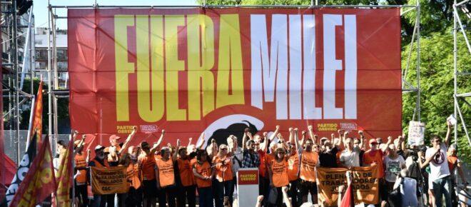 Fotografía de un acto político en un escenario al aire libre. Al fondo, un gran telón rojo muestra la consigna 'FUERA MILEI' en letras grandes de color amarillo y blanco. Sobre el escenario, un grupo de aproximadamente veinte personas posa con los puños en alto. La mayoría viste pecheras de color naranja. Frente al escenario, una multitud de espaldas observa el acto, con banderas rojas visibles en los laterales. Fotografía de un acto político en un escenario al aire libre. Al fondo, un gran telón rojo muestra la consigna 'FUERA MILEI' en letras grandes de color amarillo y blanco. Sobre el escenario, un grupo de aproximadamente veinte personas posa con los puños en alto. La mayoría viste pecheras de color naranja. Frente al escenario, una multitud de espaldas observa el acto, con banderas rojas visibles en los laterales.