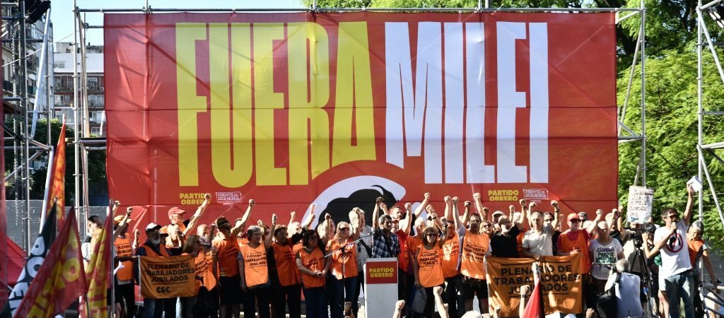 Fotografía de un acto político en un escenario al aire libre. Al fondo, un gran telón rojo muestra la consigna 'FUERA MILEI' en letras grandes de color amarillo y blanco. Sobre el escenario, un grupo de aproximadamente veinte personas posa con los puños en alto. La mayoría viste pecheras de color naranja. Frente al escenario, una multitud de espaldas observa el acto, con banderas rojas visibles en los laterales. Fotografía de un acto político en un escenario al aire libre. Al fondo, un gran telón rojo muestra la consigna 'FUERA MILEI' en letras grandes de color amarillo y blanco. Sobre el escenario, un grupo de aproximadamente veinte personas posa con los puños en alto. La mayoría viste pecheras de color naranja. Frente al escenario, una multitud de espaldas observa el acto, con banderas rojas visibles en los laterales.