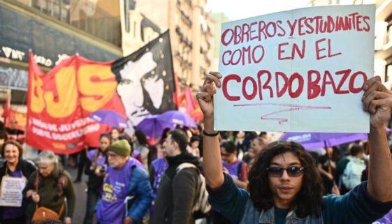 "Un joven sostiene en alto un cartel escrito a mano durante la cuarta marcha federal univeristaria. El texto en letras rojas dice: 'Obreros y estudiantes como en el Cordobazo'. Al fondo se observan banderas rojas y negras, paraguas violetas y una multitud de manifestantes." "Un joven sostiene en alto un cartel escrito a mano durante la cuarta marcha federal univeristaria. El texto en letras rojas dice: 'Obreros y estudiantes como en el Cordobazo'. Al fondo se observan banderas rojas y negras, paraguas violetas y una multitud de manifestantes."