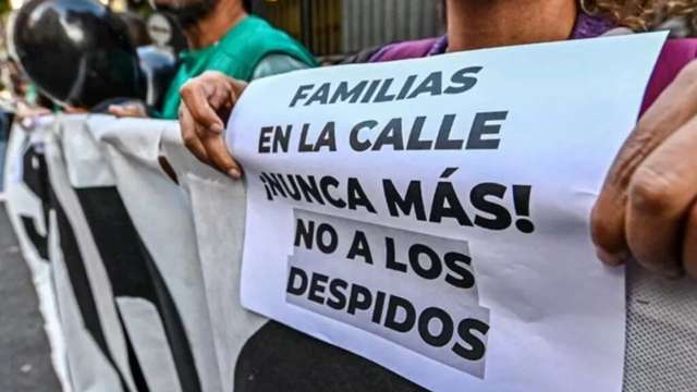 "Primer plano de una persona sosteniendo un cartel blanco impreso en español durante una manifestación callejera. El texto dice: 'FAMILIAS EN LA CALLE ¡NUNCA MÁS! NO A LOS DESPIDOS'. Las manos de la persona sujetan el cartel por ambos lados. Al fondo, borrosos, se distinguen otros manifestantes y el contorno de un casco de motocicleta."