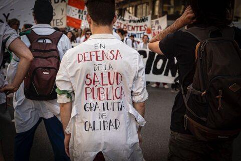 "Un trabajador de salud pública con su uniforme puesto en una movilización. Se encuentra de espaldas, donde se encuentra escrito 'en defensa de la salud publica, gratuita, de calidad' de fondo hay mas personas y pancartas" "Un trabajador de salud pública con su uniforme puesto en una movilización. Se encuentra de espaldas, donde se encuentra escrito 'en defensa de la salud publica, gratuita, de calidad' de fondo hay mas personas y pancartas"