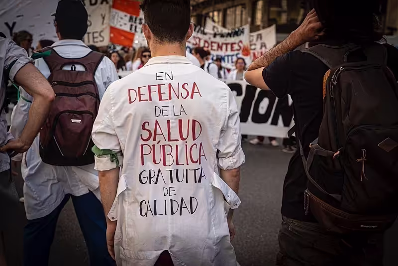 "Un trabajador de salud pública con su uniforme puesto en una movilización. Se encuentra de espaldas, donde se encuentra escrito 'en defensa de la salud publica, gratuita, de calidad' de fondo hay mas personas y pancartas" "Un trabajador de salud pública con su uniforme puesto en una movilización. Se encuentra de espaldas, donde se encuentra escrito 'en defensa de la salud publica, gratuita, de calidad' de fondo hay mas personas y pancartas"